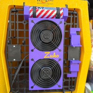 Pu&ograve; includere: Sistema di ventilazione viola con due ventole e un cavo di alimentazione. Le ventole sono montate su un pannello viola con la parola "Zuko" scritta in giallo. Il sistema &egrave; attaccato a una gabbia per cani gialla con il testo "Ruff Land Performance Kennels" e "PK9 Emergency Information" visibile.