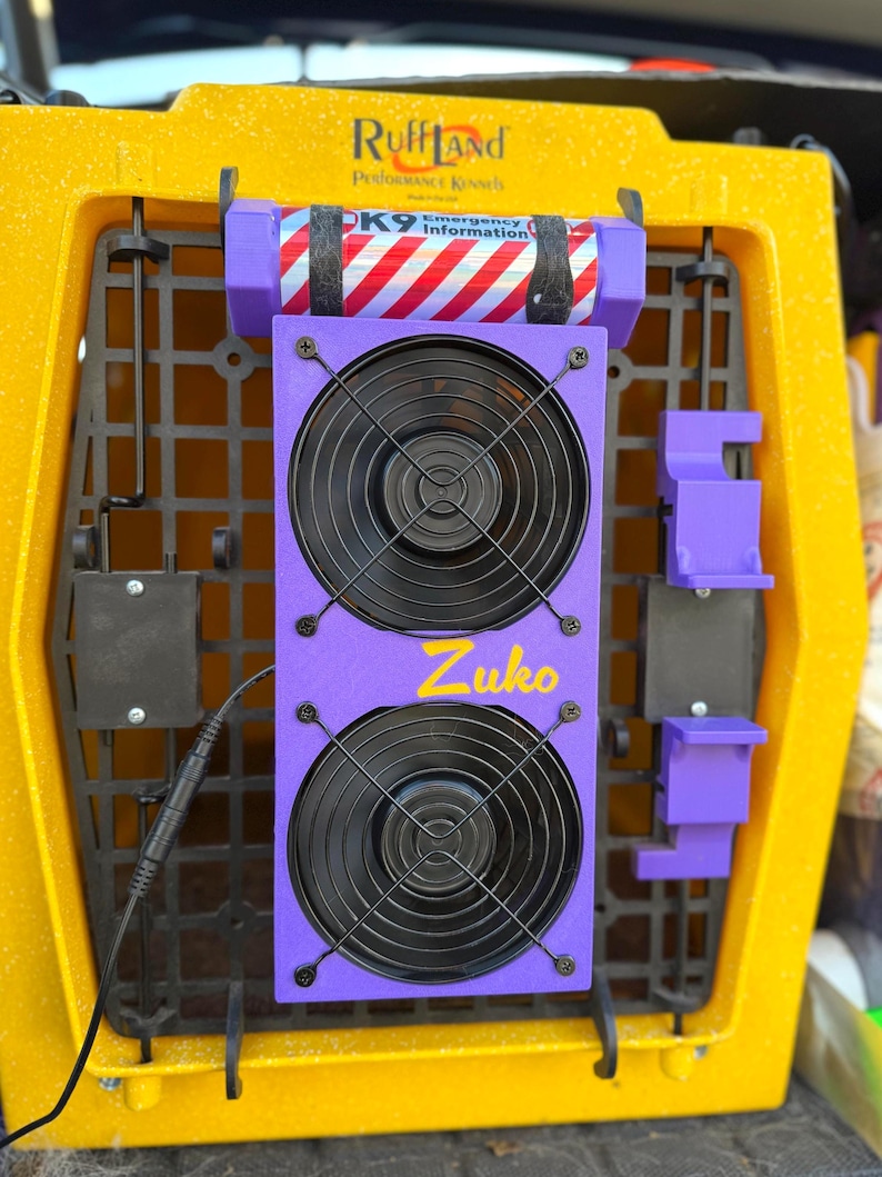 Pu&ograve; includere: Sistema di ventilazione viola con due ventole e un cavo di alimentazione. Le ventole sono montate su un pannello viola con la parola "Zuko" scritta in giallo. Il sistema &egrave; attaccato a una gabbia per cani gialla con il testo "Ruff Land Performance Kennels" e "PK9 Emergency Information" visibile.