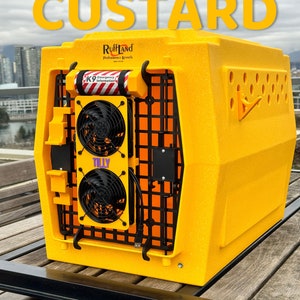 May include: A yellow Ruffland Performance Kennels dog crate with a black metal door and two black fans. The crate has a white sticker with the text "K9 Emergency Information" and a purple sticker with the text "Tilly". The crate is sitting on a wooden table.