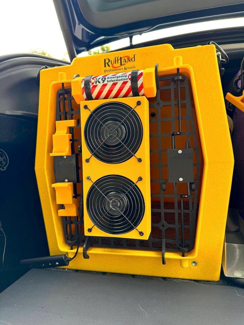 Dual dog crate fan with custard yellow 3D-printed housing, securely mounted to the door of a Ruffland Performance Kennel using bolt-on hardware. Features two enclosed cooling fans for optimal airflow, customized with a dog’s name, and installed in a vehicle for travel. Also shown are mounted accessories including a K9 emergency information canister. Ideal for keeping dogs cool and safe during transport or daily crate use.
