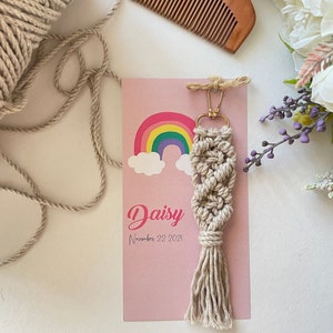 Op de afbeelding: Een beige macramé sleutelhanger met een goudkleurige sluiting en een kwast. De sleutelhanger wordt getoond op een roze kaart met een regenboogafbeelding en de naam "Daisy" en de datum "22 november 2021". Een houten kam en bloemen staan op de achtergrond.