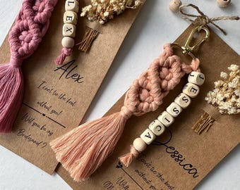 May include: Two pink and white macrame keychains with wooden beads spelling out the names "Alex" and "Jessica". Each keychain is attached to a brown card with a message asking "Will you be my Bridesmaid?" and "Will you be my Maid of Honor?"