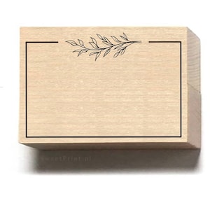 May include: A wooden stamp with a rectangular frame and a leafy branch design at the top. The stamp is blank inside the frame.