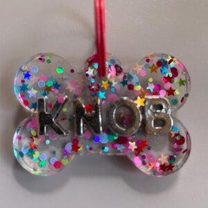 May include: A clear, bone-shaped pet tag made of resin, embedded with multicoloured star and circular glitter. The word KNOB is spelled out in silver, metallic, three-dimensional capital letters across the centre. A pink plastic ring is attached.