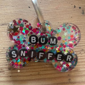 Könnte beinhalten: Eine knochenförmige Haustiermarke aus klarem Harz, gefüllt mit buntem Glitzer und kleinen sternförmigen Pailletten. Die Marke enthält schwarze runde Perlen mit weißen Buchstaben, die den Text BUM SNIFFER bilden, und einen silbernen Metallring.