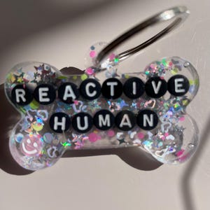 Könnte beinhalten: Ein knochenförmiger Schlüsselanhänger aus Kunstharz mit dem Text REACTIVE HUMAN in schwarzen Buchstabenperlen. Das klare Harz ist mit buntem, holografischem Stern- und Herzglitzer sowie einem kleinen rosa Smiley-Anhänger und einem Metallring gefüllt.