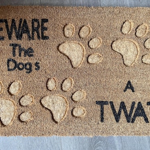 May include: A brown coir doormat with the text "Beware the Dogs A Twat" in black lettering. The doormat has several paw prints embossed into the surface.