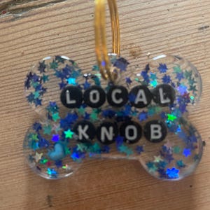 Könnte beinhalten: Eine knochenförmige Haustiermarke aus klarem Harz, gefüllt mit blauem und türkisfarbenem sternförmigem Glitzer. Die Marke enthält schwarze runde Perlen mit weißen Buchstaben, die den Text LOCAL KNOB bilden, sowie einen goldenen Metallring.