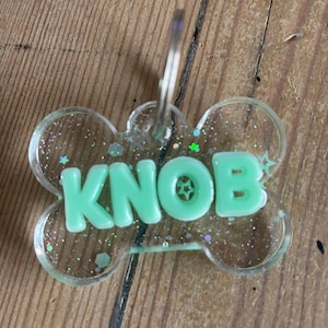 May include: A clear, bone-shaped pet tag with the name "KNOB" in mint green, glitter accents, and a silver keyring. The tag is made of clear acrylic and is designed for a pet.