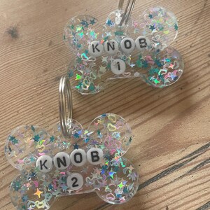 May include: Two clear, bone-shaped resin tags featuring embedded iridescent star and heart-shaped glitter. Each tag includes a silver metal split ring and white letter beads spelling KNOB, with a single number bead below reading 1 or 2.