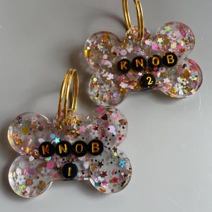 May include: Two bone-shaped resin tags featuring a clear base filled with multicolored glitter, including small stars, hearts, and flowers. Each tag has a gold-tone metal ring and black beads spelling KNOB 1 and KNOB 2 respectively.