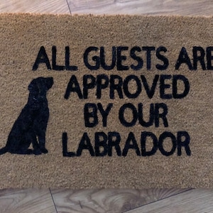 May include: A brown doormat with the text "All guests are approved by our Labrador" in black lettering. A black silhouette of a Labrador Retriever is sitting to the left of the text.