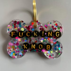 May include: A clear, bone-shaped resin pet tag filled with multicoloured star and circle glitter. The tag features black circular beads with gold lettering spelling out the phrase FUCKING KNOB in two rows, attached to a gold-coloured metal ring.