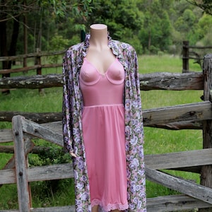 以下が含まれることがあります： マネキンのヴィンテージピンクのランジェリーセット。レーストリム付きのスリップと、お揃いのローブが含まれています。ローブは、ピンクの背景に紫と白の花のフローラルパターンが特徴です。マネキンは木のフェンスの前に立っています。