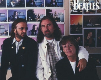 May include: A promotional image featuring three members of The Beatles. The band members are posed in front of a wall of framed images. The Beatles logo is visible in the upper right corner. The band members are wearing jackets and shirts.