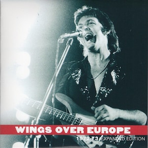Puede incluir: Portada del álbum "Wings Over Europe 1972-73 Expanded Edition". La imagen es una foto en blanco y negro de un músico cantando a un micrófono mientras toca la guitarra. El título del álbum está en texto blanco sobre una franja roja.