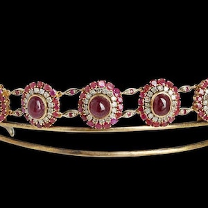 May include: A gold tiara featuring a row of red gemstones encircled by smaller diamonds. The tiara is crafted from metal and has a delicate design.
