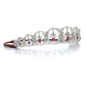 Könnte beinhalten: Eine silberne Tiara mit einem filigranen Design, das mit Rubinen und Diamanten besetzt ist. Die Tiara liegt auf einem braunen Samtkissen.