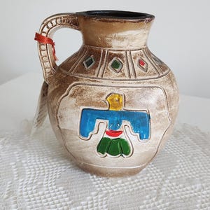 Op de afbeelding: Een bruin keramieken kruik met een handvat, met een gestileerd vogelontwerp in blauw, geel en groen. De kruik heeft een donkere rand en decoratieve ruitvormen. Een rood lint is aan het handvat gebonden.