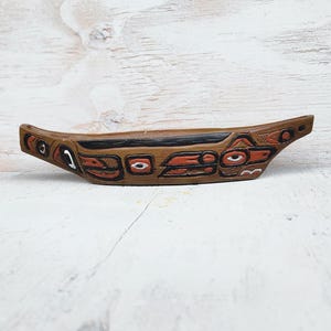 Puede incluir: Un cuenco de madera tallada marrón con diseños geométricos rojos, negros y blancos. El cuenco tiene forma curva y es probablemente un objeto decorativo o ceremonial. La obra de arte parece inspirada en el arte indígena.