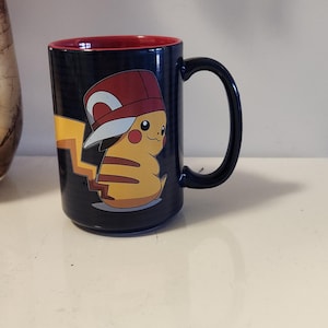 May include: A black ceramic mug with a red rim features a cartoon image of Pikachu wearing a red and white baseball cap. The mug has a white interior and a black handle.