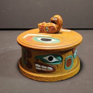 Peut inclure: Une boîte ronde en bois avec un oiseau sculpté sur le couvercle. La boîte est décorée d'un motif traditionnel amérindien dans des tons de brun, vert, rouge et blanc.