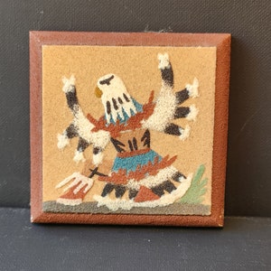 Puede incluir: Una pintura de arena enmarcada que representa a un bailarín nativo americano con plumas de águila y una falda colorida. La pintura está sobre un fondo beige y enmarcada en marrón.