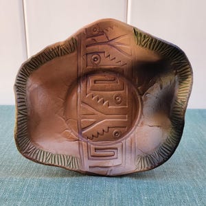 May include: A brown ceramic dish with a green rim and a geometric design. The design is carved into the surface of the dish and features a repeating pattern of triangles and circles.