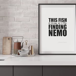 May include: A framed print with the quote "THIS FISH IS SO RAW, HE'S STILL FINDING NEMO" in bold black letters. The print is in a black frame and is displayed on a white countertop with kitchen accessories.