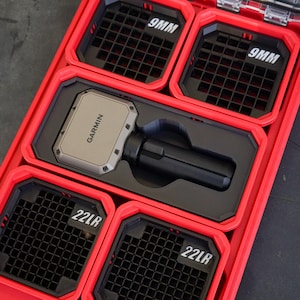 Puede incluir: Una caja de almacenamiento roja compartimentada con inserciones negras. La caja contiene un dispositivo Garmin plateado y un objeto cilíndrico negro. Los compartimentos están etiquetados "9MM" y "22LR" en texto blanco.