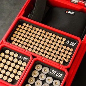 May include: A red ammunition case, open to reveal several compartments filled with different types of cartridges. The case contains 5.56 rounds, 6 cm rounds, and 20 GA rounds. A black pouch with the text "FIRST" is also visible.