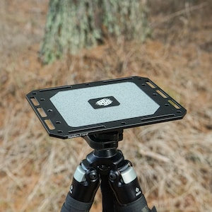 May include: A black and gray camera tripod with a rectangular top plate. The top plate has a textured gray surface and the text "Matthew 7: 24-27". The tripod is set up outdoors.