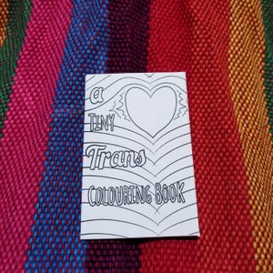 May include: A black and white colouring book with the title "A Tiny Trans Colouring Book". The cover features a heart with a wavy design and the word "Trans" in a stylised font.