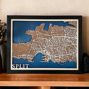 Peut inclure: Une carte en bois encadrée de Split, en Croatie, avec un fond bleu et des lignes blanches représentant les rues de la ville. Le mot "SPLIT" est écrit en lettres blanches en bas de la carte.