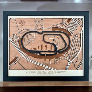 May include: Wooden laser-cut map of the Charlotte Motor Speedway in Concord, North Carolina, USA. The map features the track layout in black, with surrounding roads and buildings in a light wood colour, framed in a dark blue frame.