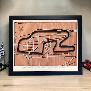 Pode incluir: Um mapa de madeira emoldurado do Circuito Internacional de Watkins Glen, com um contorno de pista preto e detalhes de estrada brancos. O mapa inclui o texto "Watkins Glen International Circuit" e uma rosa dos ventos. A moldura é preta.