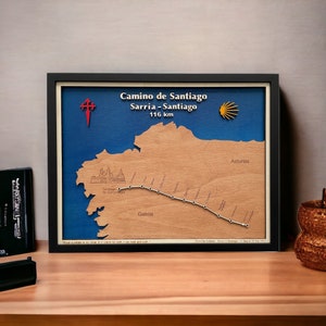 Può includere: Mappa in legno incorniciata del percorso di pellegrinaggio del Cammino di Santiago, Sarria a Santiago, 116 km. La mappa presenta un ritaglio in legno marrone chiaro della regione, uno sfondo blu e una cornice nera. Testo: Camino de Santiago, Sarria - Santiago.