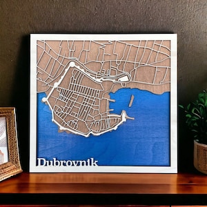 May include: Wooden map art of Dubrovnik, Croatia, with a white frame. The map features a detailed street layout in a light brown color, set against a blue sea. The city's name, "Dubrovnik," is displayed in white at the bottom.