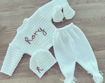 May include: A white knitted baby outfit set, including a long-sleeved top with "Rory" embroidered in brown, matching pants, a beanie with an embroidered "R", and mittens. The set is made of a textured knit fabric.