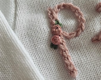 May include: A white knit sweater featuring the letter P embroidered in a dusty pink yarn. The letter is decorated with two small, dimensional pink rosebuds and tiny green leaves stitched directly onto the surface of the knitted fabric.