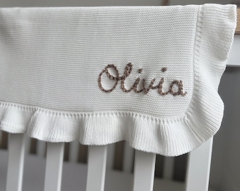 May include: A white knitted baby blanket with a ruffled edge. The name "Olivia" is embroidered in brown thread on the blanket. The blanket is draped over a white crib.