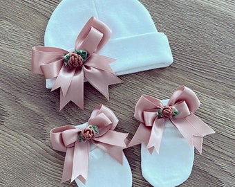 May include: A white beanie and a pair of matching white mittens, each featuring a dusty pink satin ribbon bow. Centered on each bow is a small, embroidered dusty pink rosebud with green leaves, providing a decorative accent to the set.