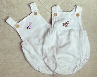 May include: Two white baby onesies with embroidered letters. The onesie on the left has a purple "E" with floral accents. The onesie on the right has a brown "E" with floral accents.