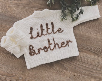 May include: A white knitted baby sweater and matching booties. The words "Little Brother" are embroidered in brown thread on the front of the sweater. The items are displayed on a wooden surface with greenery in the background.