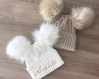 May include: Two knit beanies with double faux fur pom-poms. One beanie is white with the name Archie embroidered in light brown script, and the other is a solid beige. Both feature a cable knit pattern on the crown and a ribbed cuff at the base.