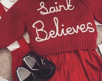 May include: A red knitted baby outfit with the words "Saint Believes" in white script. The outfit includes a sweater, shorts, a collared shirt, and black patent leather shoes. A white hanger with the name "Miranda" is also visible.