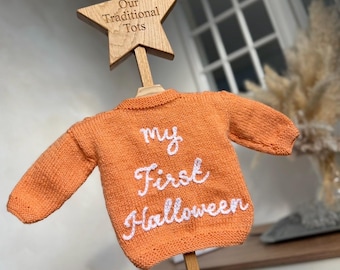 May include: An orange knitted sweater featuring the white embroidered text "my First Halloween" on the front. The sweater hangs on a wooden hanger topped with a star-shaped wooden plaque engraved with the text "Our Traditional Tots".