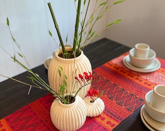 Op de afbeelding: Drie witte keramische vazen met een geribbelde textuur, gerangschikt op een rode en oranje gestreepte tafelrunner. De vazen zijn gevuld met groene bamboestokken en rode bloemen.