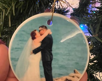 Peut inclure: Une boule de Noël ronde en céramique blanche avec une photo d'un couple s'embrassant devant un fond d'eau bleu. La boule est munie d'une ficelle dorée attachée au sommet.
