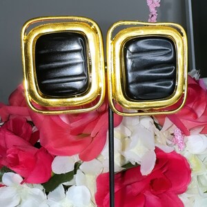 May include: A pair of gold-tone square earrings with black rectangular inserts. The earrings are displayed on a black stand against a background of pink and white flowers.